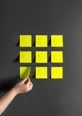 Yellow paper stickers on a dark gray wall. A hand peels off a post-it note.の写真素材
