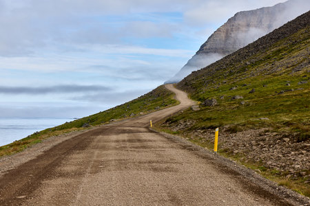 Winding gravel road along Iceland's coastal mountainside with yellow markers. Dramatic misty cliffs and ocean view create stunning scenic route.の写真素材