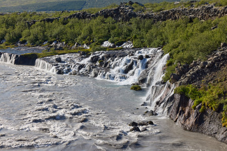 A picturesque waterfall cascades over rocky cliffs surrounded by lush greenery. The crystal clear...