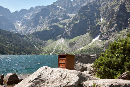 A wooden cabinet stands alone on rocky terrain near a tranquil lake. Majestic mountains rise in the background, under clear blue skies, creating a striking contrastの写真素材