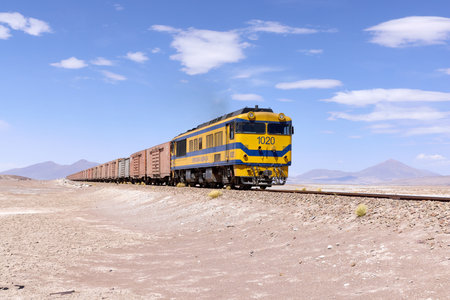 A railway locomotive pulling cargo train through Bolivian desert with distant volcanic mountains under bright blue sky with scattered clouds. 15/10/2023, Boliviaの写真素材