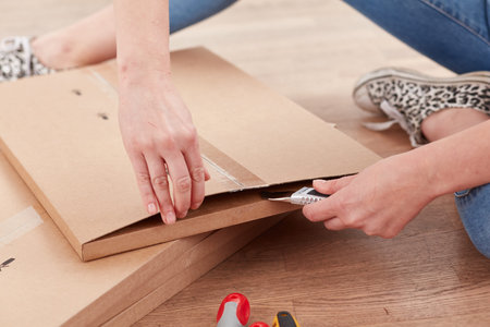 A person is seated on the floor, carefully using a utility knife to cut open cardboard boxes. They are preparing to unpack their new belongings in a bright indoor spaceの写真素材