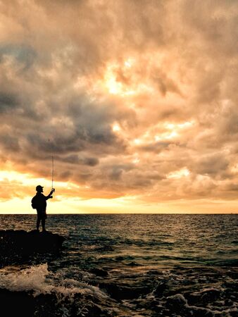Fisherman on rock watching the sunset reddish colors in the backgroundの写真素材