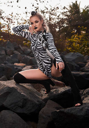 a girl poses on rocks in a zebra swimsuit with black lips, black high boots and a long braidの写真素材