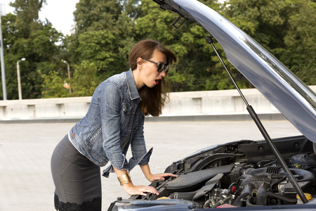 Confused young woman looking at broken down car engine on streetの写真素材