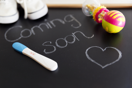 Waiting for labour, maternity concept. "Coming soon" words on black chalkboard with positive pregnancy test, white baby shoes and rattleの写真素材