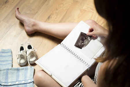 Woman placing baby's sonogram into baby's first year memory book. Baby clothes and sneakers laying on the floorの写真素材