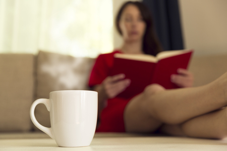 Woman with cup of hot tea reading book by window on sofa at home. Focus on cup of teaの写真素材