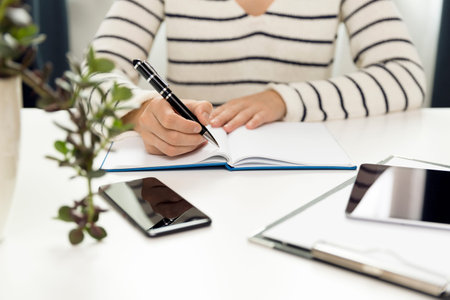 Young business woman sitting at table and writing in notebook. On table is smartphone, and tablet....