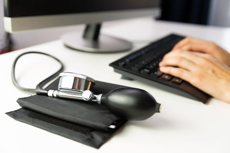 Blood pressure gauge on a white table, doctor typing on a keyboard in background.