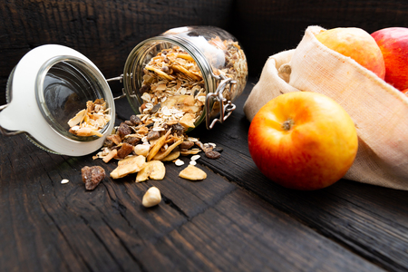 Homemade granola in glass jar and apples in reusable fabric bag on dark wooden table with texture. Healthy breakfast concept and zero waste conceptの写真素材