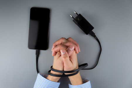 Female hands and smartphone. Woman's hands trapped and wrapped on wrists with mobile phone cable as handcuffs. Addiction to internet and social networksの写真素材