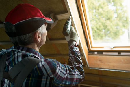 Attic insulation and renovation. Man fixing metal frame using an electric screwdriver on ceiling covered with rock woolの写真素材