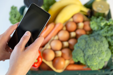 Food delivery service - woman holding smartphone in front of the box of groceriesの写真素材
