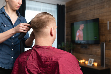 Wife cutting husband's hair at home in front of TV with clipper and holding comb in other hand.