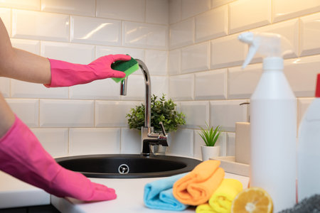 Womens hands cleaning kitchen tap on a black sink with rubber gloves and in front blurred cleaning...