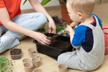 Home hobbies gardening. Young mother teaching son botany by pooring soil in pots for seeds.
