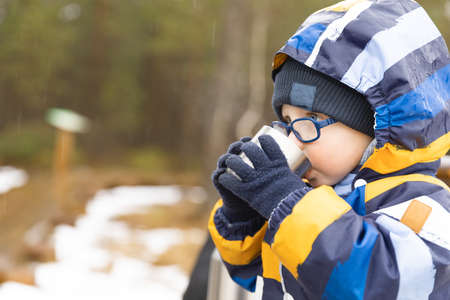 Young boy drinks hot beverage from a cup in a cold raini day. Resting, taking a break during hiking. Healthy living concept.の写真素材