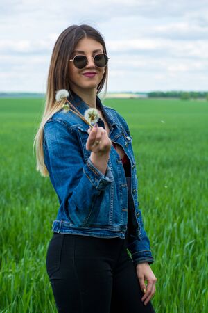 A girl in jeans clothes with beautiful hair stands on a green field with a dreamy look on her face, holds a dandelion in her hand. Warm spring dayの写真素材