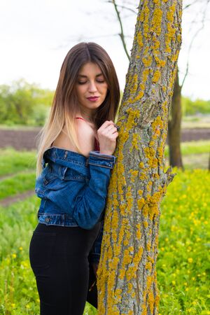 A girl in denim clothes with beautiful hair stands next to a tree. Warm spring dayの写真素材