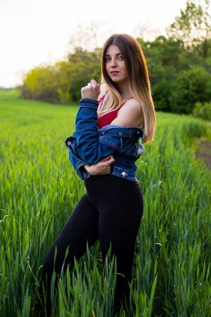 A girl in denim clothes with beautiful hair stands on a green field with a dreamy look on her face. Warm spring dayの写真素材