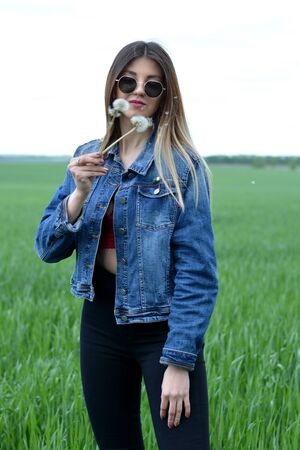 A girl in jeans clothes with beautiful hair stands on a green field with a dreamy look on her face, holds a dandelion in her hand. Warm spring dayの写真素材