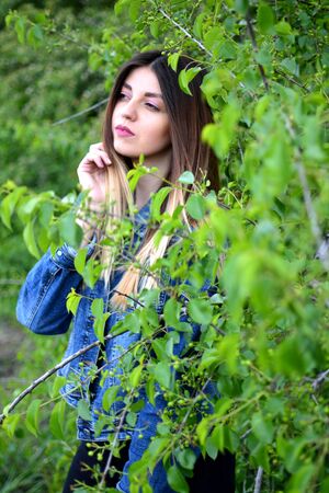 Next to the branches of a tree is a girl in jeans clothes with beautiful hair with a dreamy look on her face. Warm spring dayの写真素材