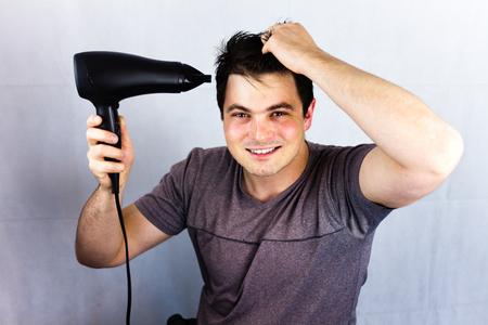Teenager smiling while he dries his hair. Man holding hair blower. Young man dries hair with an electric fan. Preparing a haircut for going to a night lifeの写真素材