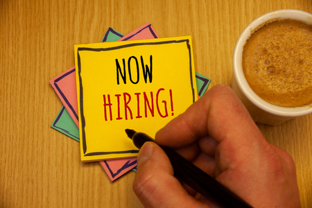 Text sign showing Now Hiring Motivational Call. Conceptual photo Workforce Wanted Employees RecruitmentMan creating yellow pink blue papers Hand holding black pen cup of coffeeの写真素材
