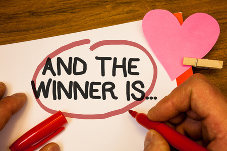 Handwriting text And The Winner Is . Concept meaning Announcing as first place in competition or race Human hand retain red pen circled black words paper hook rivet pink heartの写真素材