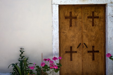 Four Crosses Mounted on Two Panel Closed Door. All Materials are made of Wood against White Cemented Wall. Plants with Pink Flowers Placed at the Entranceの写真素材