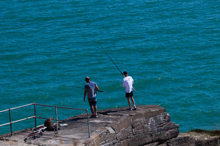 Brighton, UK - June 2018 Two Men Standing on Rock Ashore Holding Fishing Rod. Wavy Crystal Blue Water on Sunny Day. Bicycle Parked nearby Behind Railings. Watersport Activity Ideasのeditorial素材