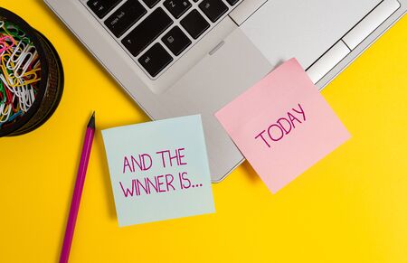 Text sign showing And The Winner Is. Business photo text announcing a demonstrating or thing that wins something Trendy metallic laptop sticky notes clips container pencil colored backgroundの写真素材