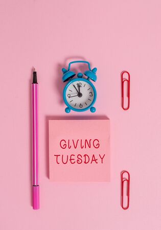 Conceptual hand writing showing Giving Tuesday. Concept meaning international day of charitable giving Hashtag activism Metal alarm clock blank notepad marker clips colored backgroundの写真素材