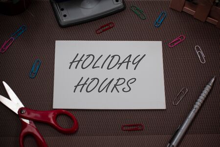 Handwriting text writing Holiday Hours. Conceptual photo employee receives twice their normal pay for all hours Scissors and writing equipments plus plain sheet above textured backdropの写真素材