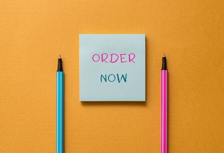 Text sign showing Order Now. Business photo showcasing the activity of asking for goods or services from a company Front view blank colored sticky note two ballpoints cool yolk backgroundの写真素材