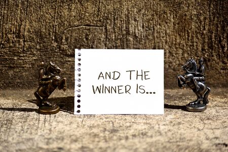 Conceptual handwriting showing And The Winner Is. Concept meaning announcing a demonstrating or thing that wins something Two Bronze Chessmen Knights Ring Notepad Sheet Folded Halfの写真素材