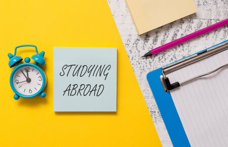 Word writing text Studying Abroad. Business photo showcasing learn outside of home in foreign country Travelling Notepads clipboard colored paper sheet marker alarm clock wooden backgroundの写真素材