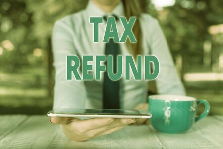 Handwriting text Tax Refund. Conceptual photo refund on tax when the tax liability is less than the tax paid Business woman sitting by the table with cup of coffee and mobile phoneの写真素材