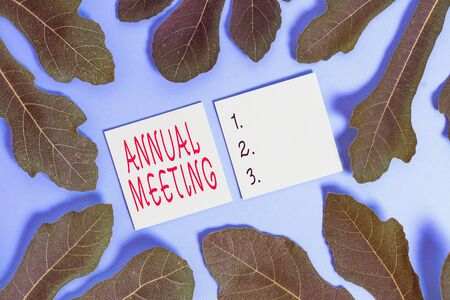 Word writing text Annual Meeting. Business photo showcasing yearly meeting of the general membership of an organization Leaves surrounding notepaper above an empty soft pastel table as backgroundの写真素材