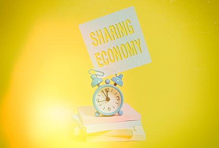 Word writing text Sharing Economy. Business photo showcasing economic model based on providing access to goods Metal alarm clock blank sticky note stacked notepads colored backgroundの写真素材