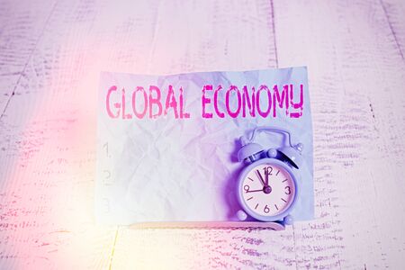 Text sign showing Global Economy. Business photo showcasing System of industry and trade around the world Capitalism Mini blue alarm clock stand tilted above buffer wire in front of notepaperの写真素材