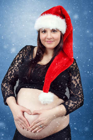 beautiful smiling pregnant young santa woman tenderly holding her tummy, hand in heart shape and with snowing in backgroundの写真素材
