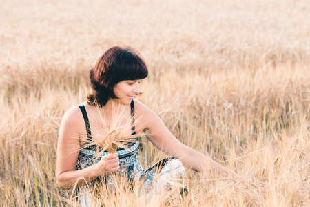 Middle aged beauty woman in a summer dress with no makeup relaxing in countryside tearing into bouquet of golden barley with her hand, summer conceptの写真素材