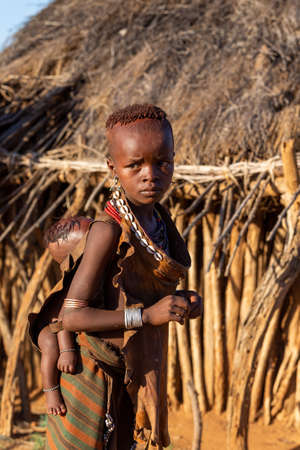 Turmi, Omo River Valley, Ethiopia - May 10, 2019: Portrait of a Hamar children with with baby on back in village. The Hamer are a primitive tribe and the women have many decorations. South Ethiopia Africaのeditorial素材