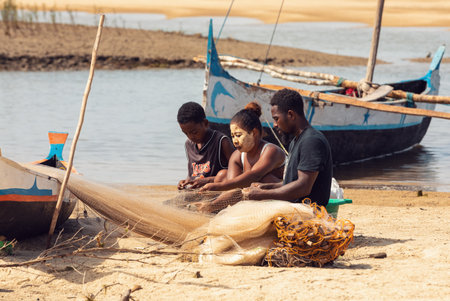 Morondava, Madagascar - November 3, 2022: Fisherman and woman repairing fishing nets at the estuaries of a river, canal Hellot. The woman has a traditionally Malagasy painted faceのeditorial素材