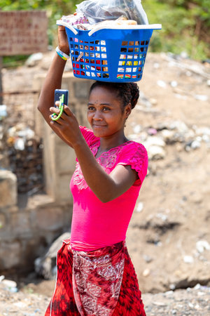 Miandrivazo, Madagascar - November 2nd, 2022: A woman carrying a basket on her head captures a photo using an old-fashioned mobile phone.のeditorial素材