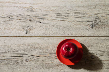 white wooden background with a red apple on a red saucer with shadowの写真素材