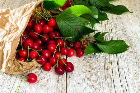 Ripe red cherry with leaves scattered from a paper bag on a white wooden table