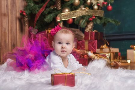 Cute baby girl wearing pink skirt and red headband, lying on white carpet near christmas trees. Christmas giftsの写真素材
